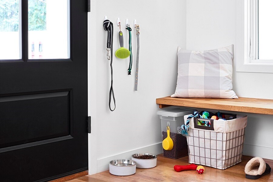 Prydlig entré med Command-krokar som håller hundkoppel, borste och halsband. Nedanför finns matskålar, en förvaringslåda och en leksakskorg. En vadderad bänk ovanför.

