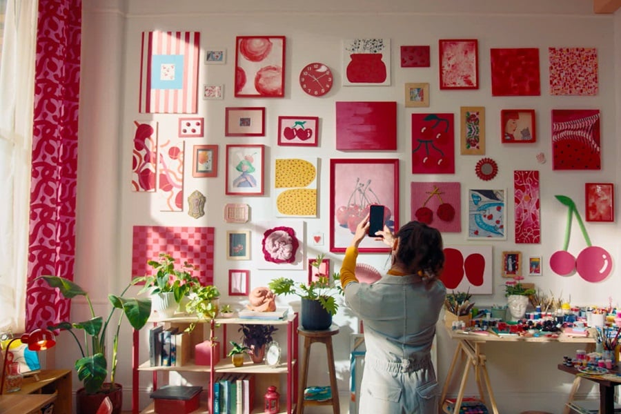 Vardagsrum med körsbärstemat konst upphängd med Command‑remsor, samt en hylla med böcker och växter och ett bord med konstmaterial.

